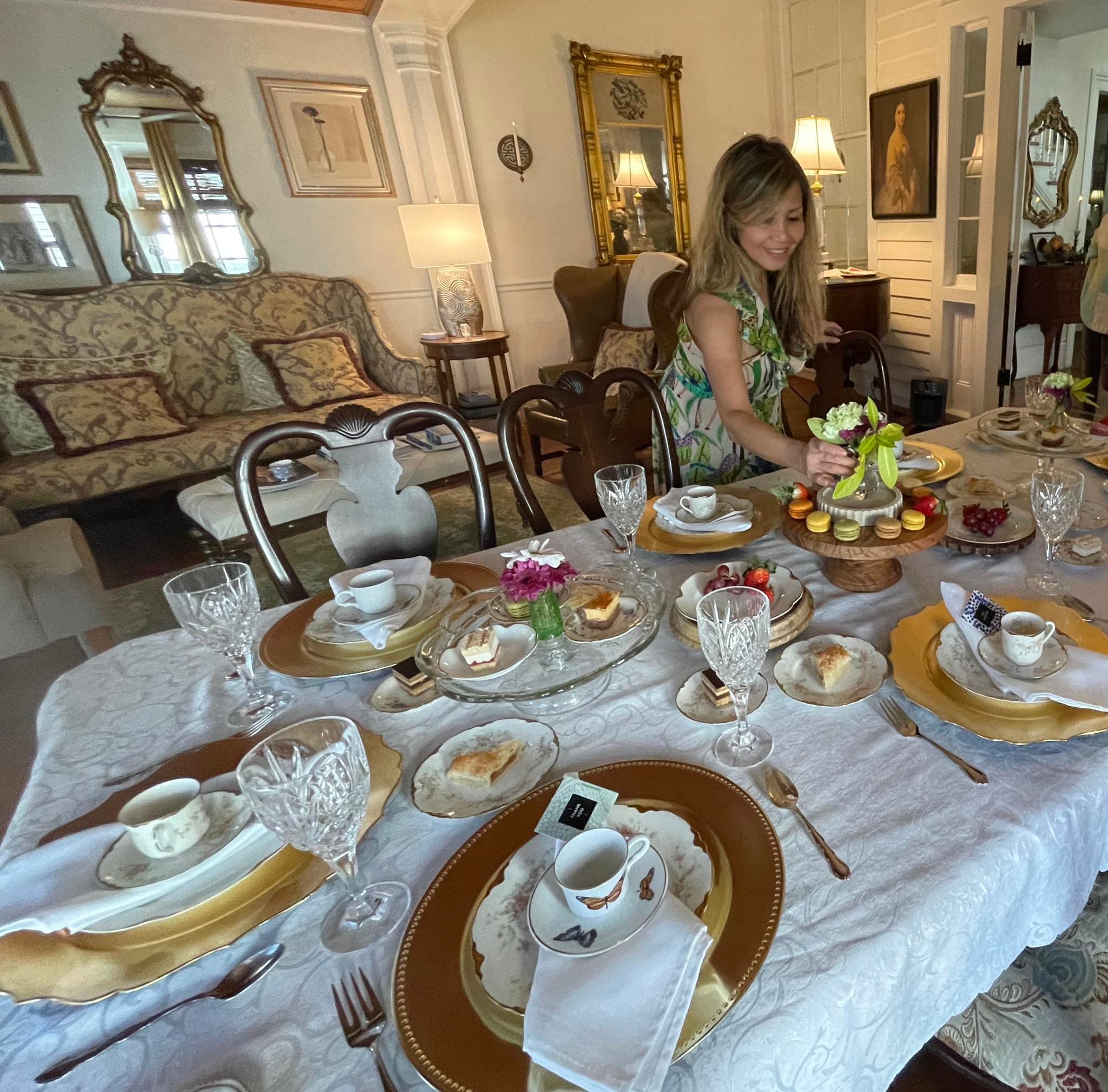 A woman arranges a beautifully set dining table with ornate dishes and floral decorations in an elegant room.
