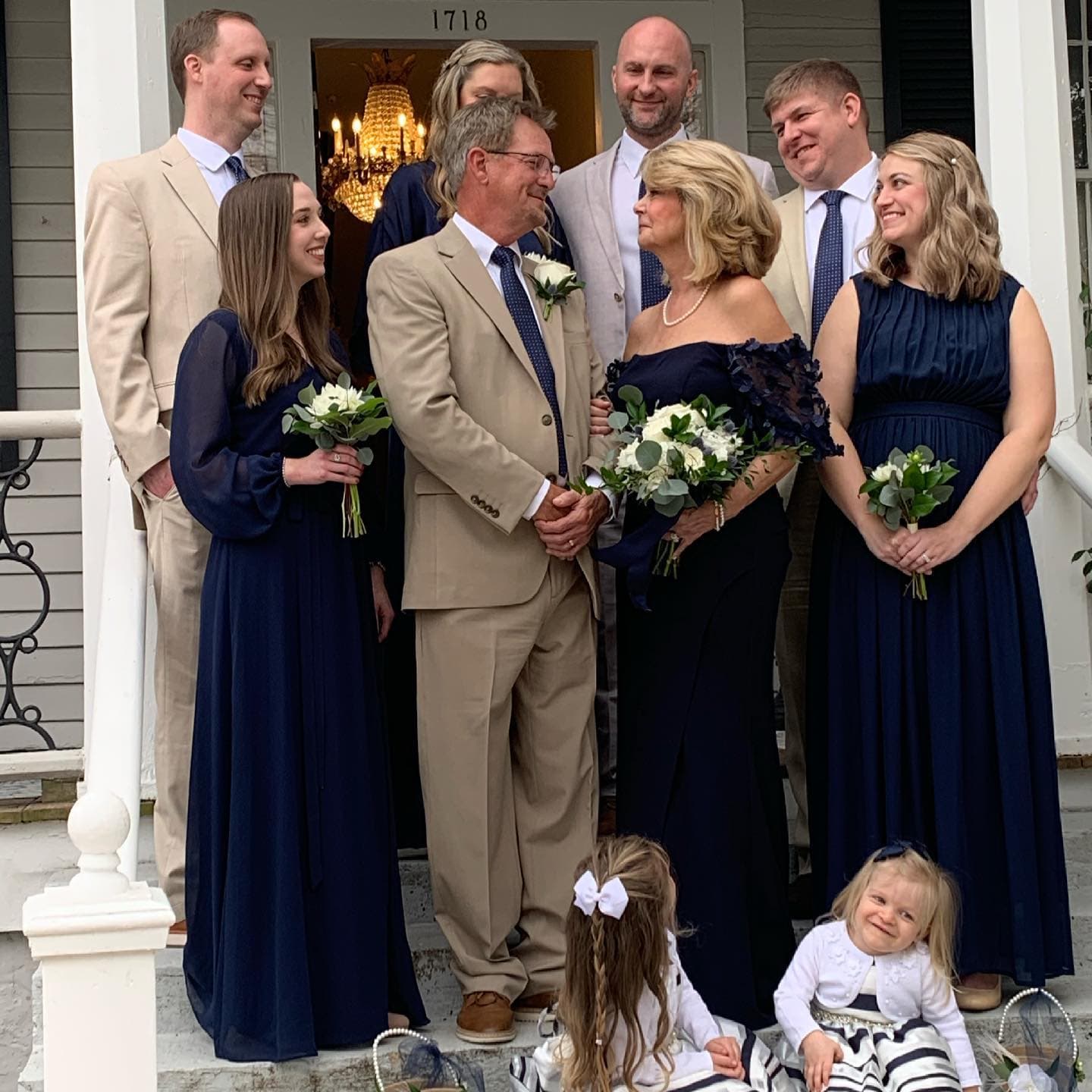 A wedding party poses on the steps of a building, dressed in formal attire, with flowers in hand and smiling at each other.