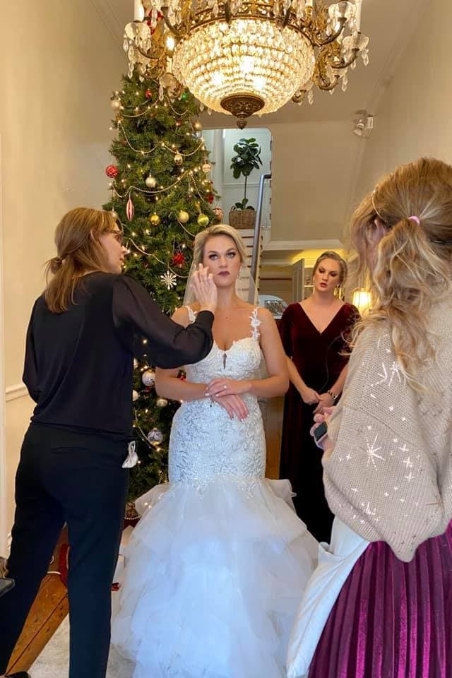 A bride is getting her makeup done while surrounded by two women in formal attire and a decorated Christmas tree in the background.