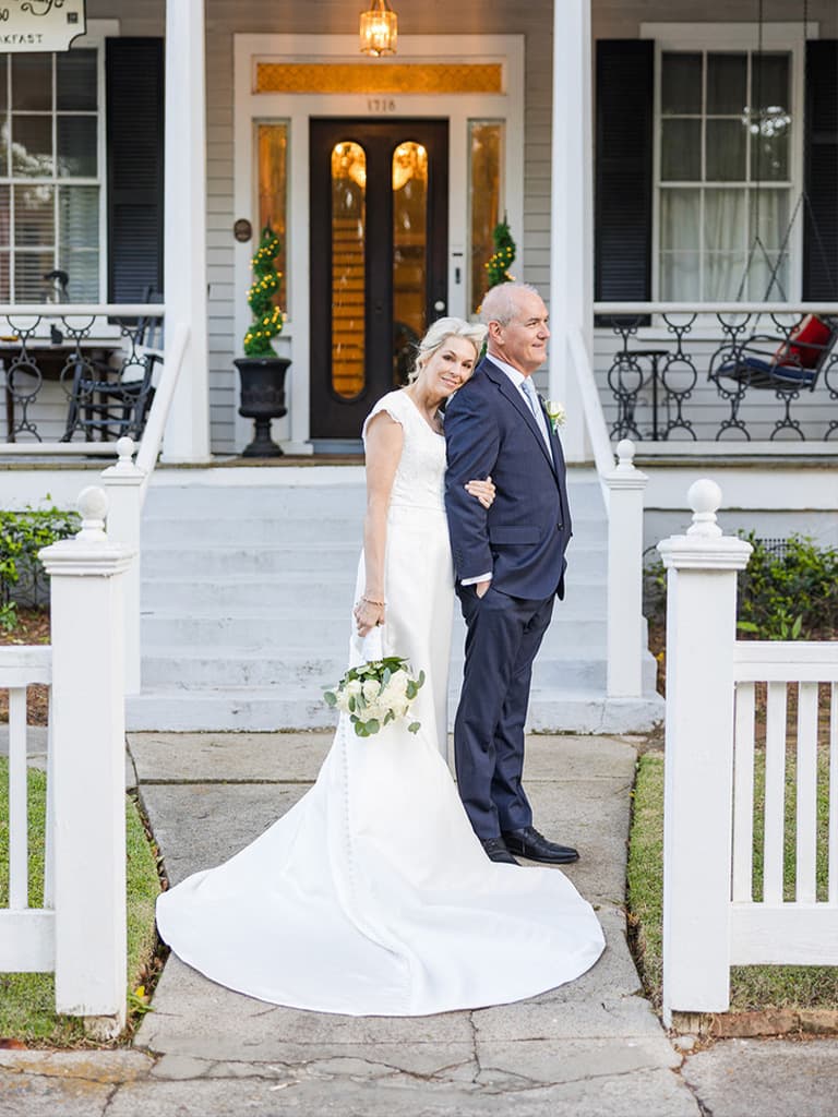 A bride and groom stand together on the steps of a house, smiling at the camera.