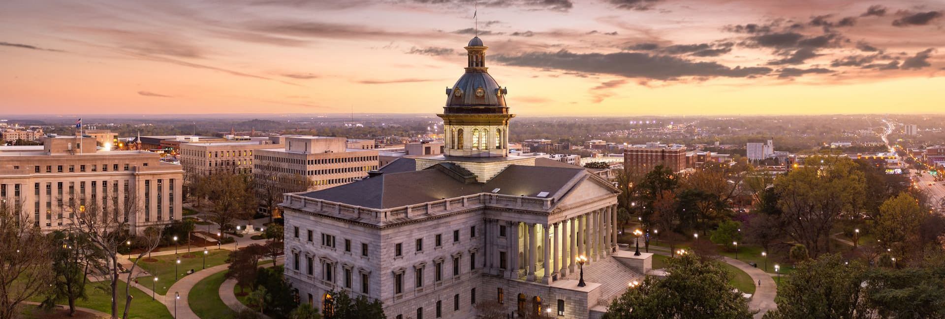 A panoramic view of a stately building with a dome at sunset, surrounded by greenery and cityscape.