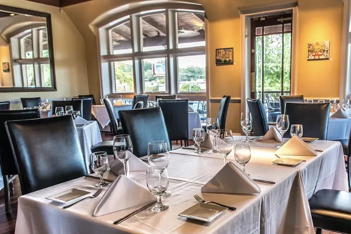 A cozy dining room with neatly set tables, elegant tableware, and large windows offering a view outside.